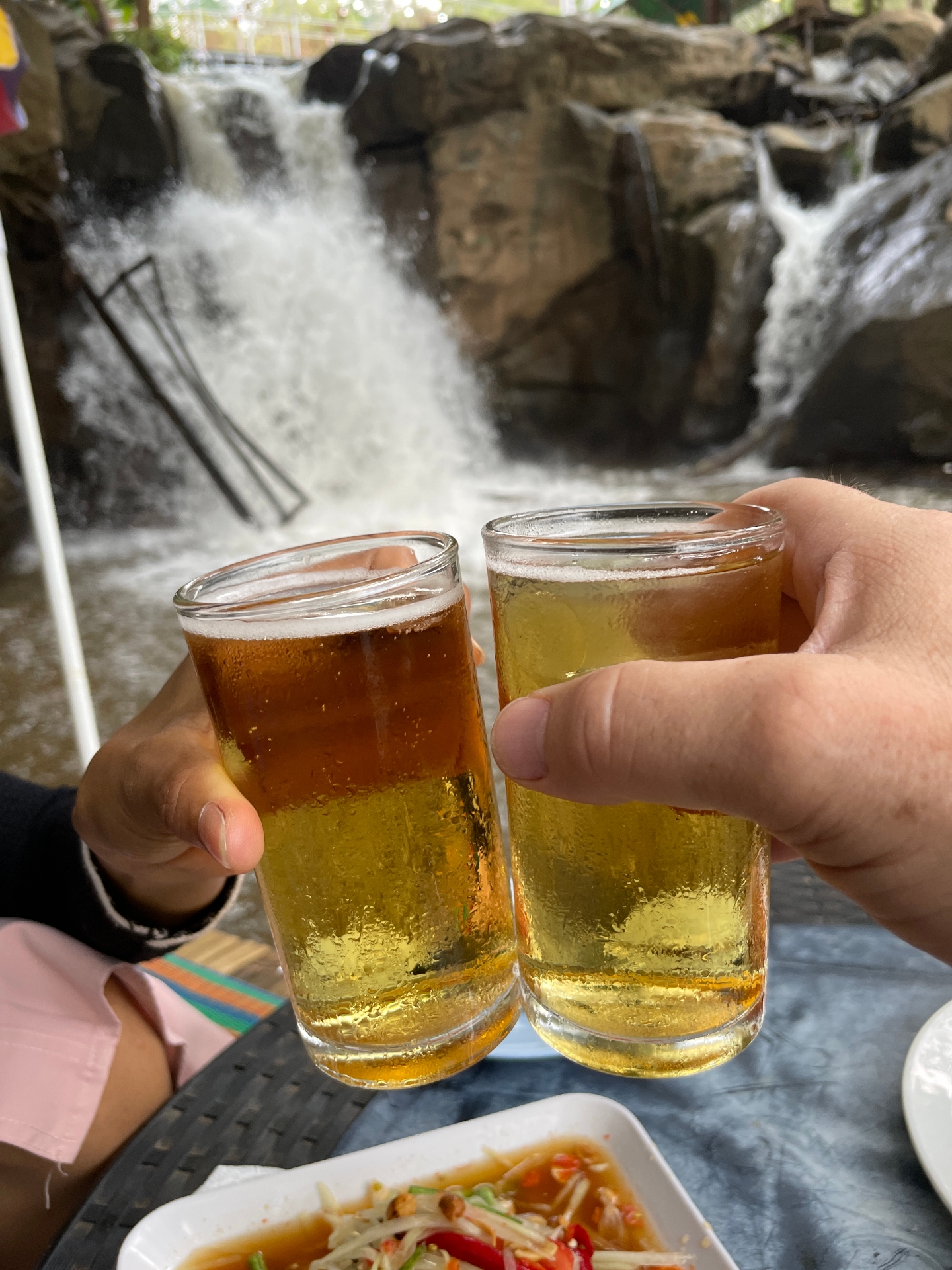 Two cold beers toasted in front of a waterfall in Thailand